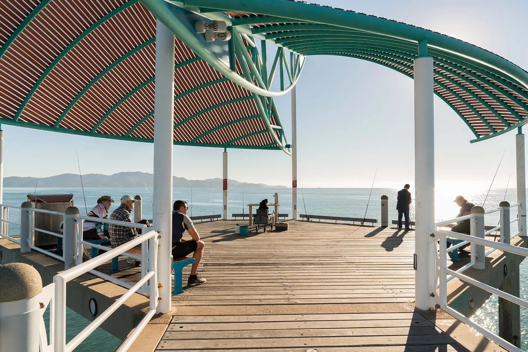 People relaxing and fishing on a shaded public jetty by the sea.